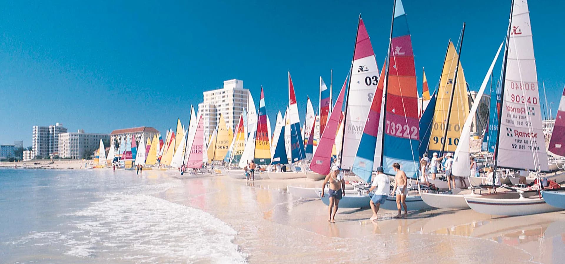 Filas de coloridas velas de catamarán sobre la arena blanca de Hobie Beach bajo un cielo azul despejado en Gqeberha.